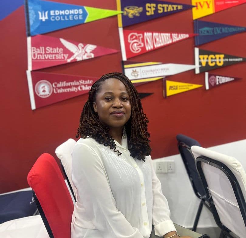 Promis Adetoro, a young woman with dreadlocks wearing a white shirt, sits in front of a wall of university flags.